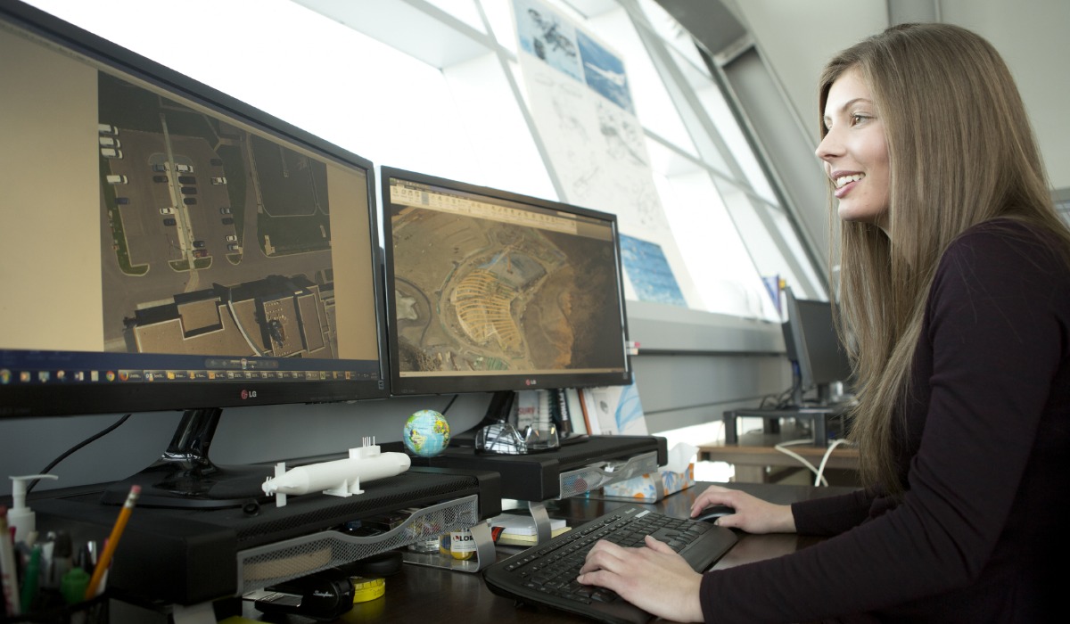 A woman working at a desk with two computer monitors displaying 3D models and maps.