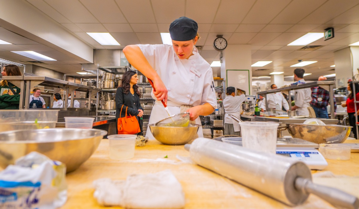 A cook apprentice wearing a chef’s uniform and hat mixes ingredients in a large metal bowl in a busy commercial kitchen filled with other people cooking and preparing food.