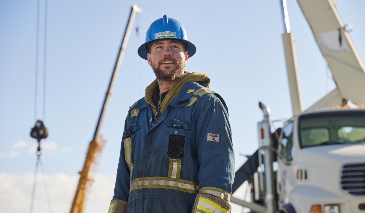 A crane operator wearing a blue hard hat and protective coveralls stands outdoors near cranes and heavy equipment, looking off into the distance under a clear sky.