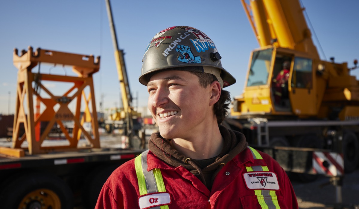 A smiling crane operator wearing a sticker-covered hard hat and red safety coveralls stands outdoors at a crane operation site with heavy machinery in the background.