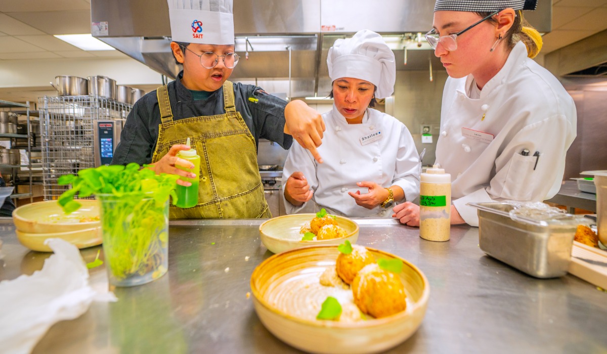 Two chefs and a culinary student collaborating in a commercial kitchen, plating a dish with garnishes.