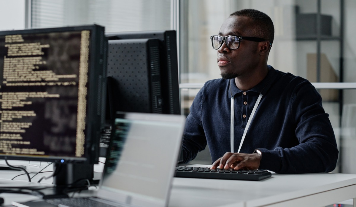 A focused cyber security analyst sitting at a desk, typing on a keyboard with multiple computer screens displaying code in the background.