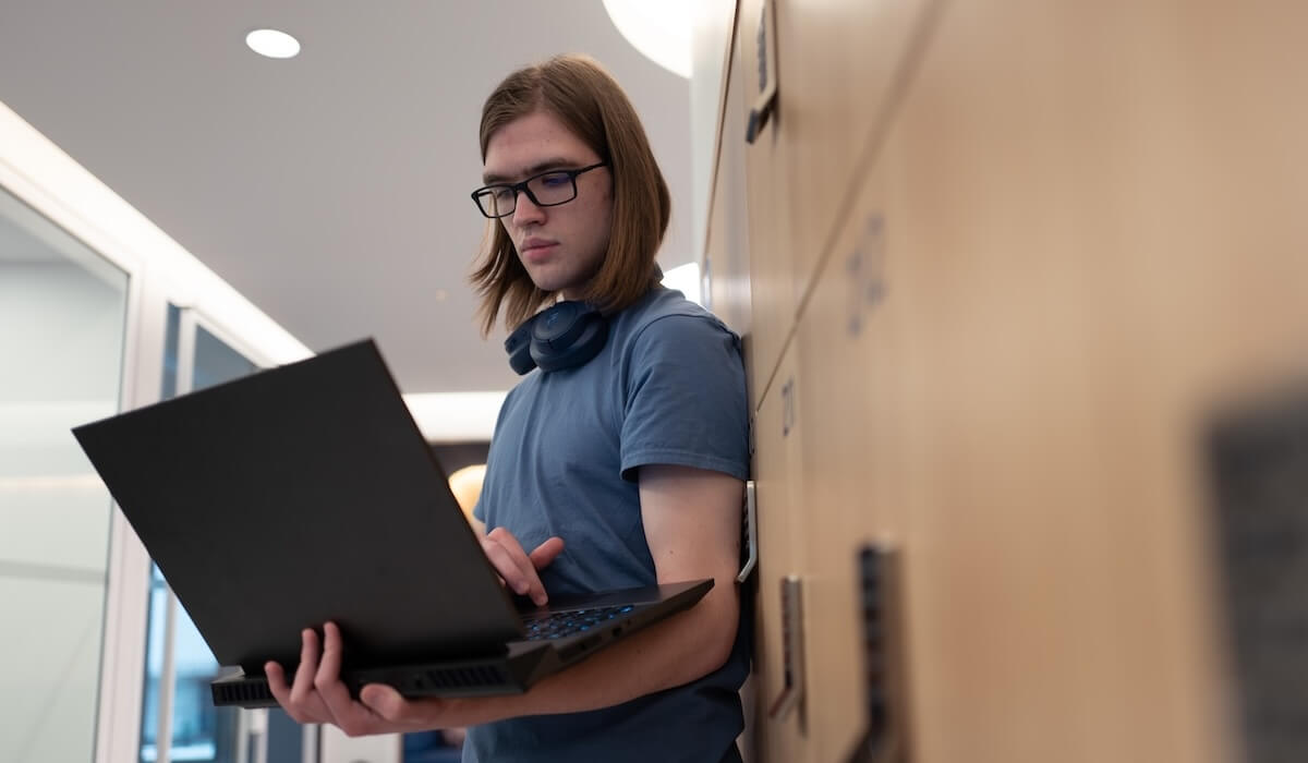A student standing in a hallway looking at a computer balanced on their arm. 