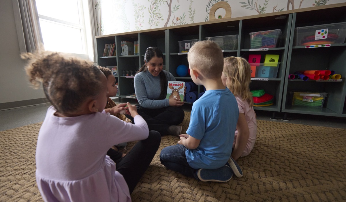 A teacher reading a book to four young children sitting on a carpet in a classroom setting.