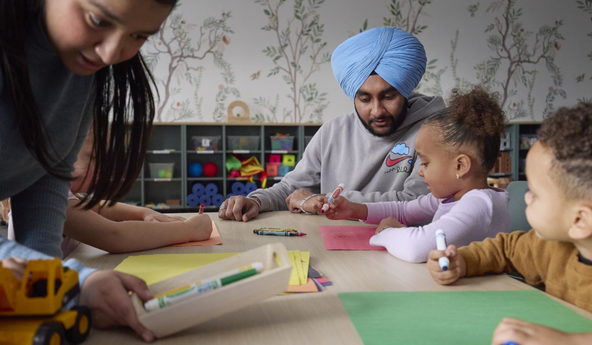 A man wearing a turban and a woman engage with children at a colorful table, where they are using art supplies for a craft activity.