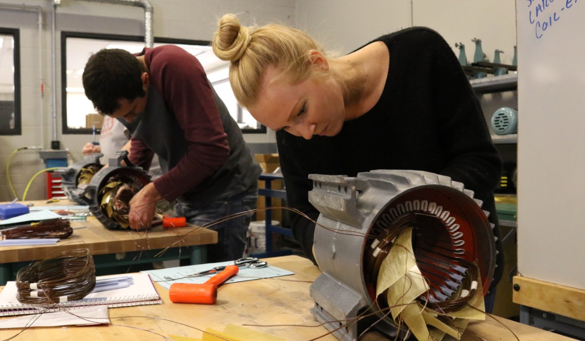 Two people work on assembling or repairing electric motor components at a workshop table, surrounded by tools, wiring, and technical notes.