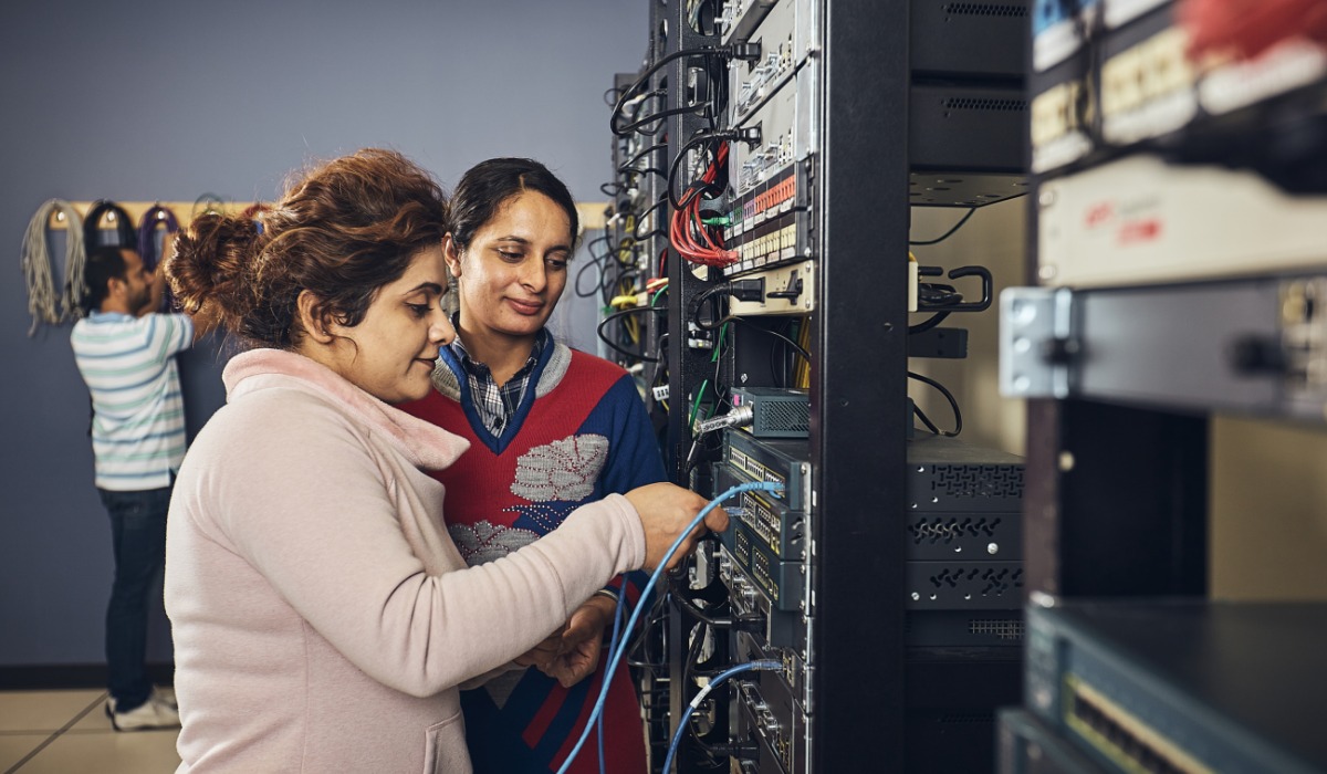 Two women working together on a network server, connecting cables while a man works in the background.