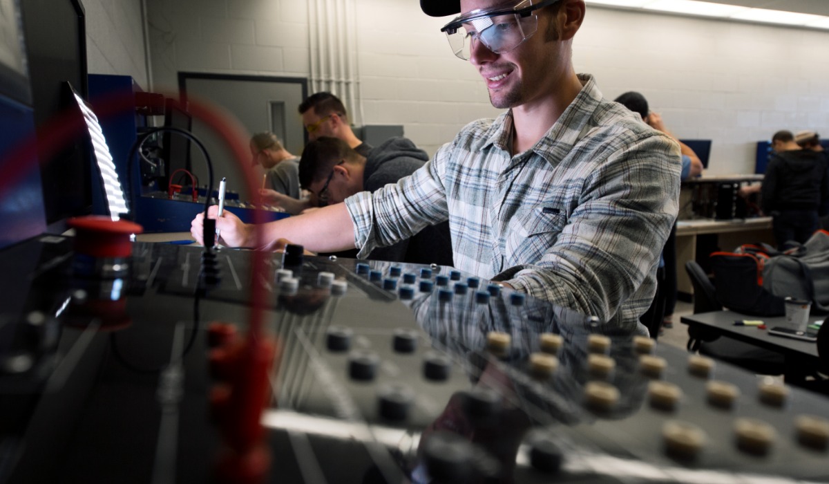 A student wearing safety glasses works with electrical panels and wiring in a classroom lab, surrounded by other students engaged in similar hands-on electrical training.