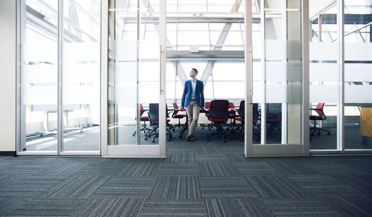 A professional man walking through a modern office corridor with glass walls and a conference room in the background.