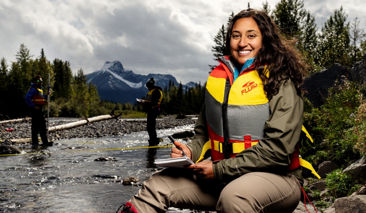 A woman in a bright life jacket smiles while taking notes by a river, with two figures performing research in the background. Mountains and a cloudy sky are visible behind her.