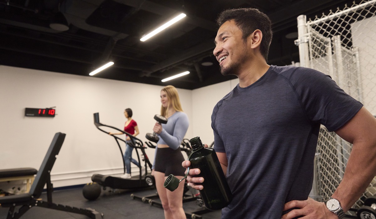 A diverse group of individuals exercising in a gym setting, with a man holding a water bottle while smiling and two others using workout equipment.