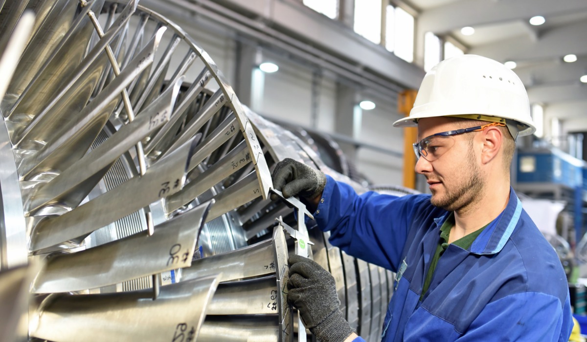 A technician in a hard hat measuring turbine blades in an air hangar.