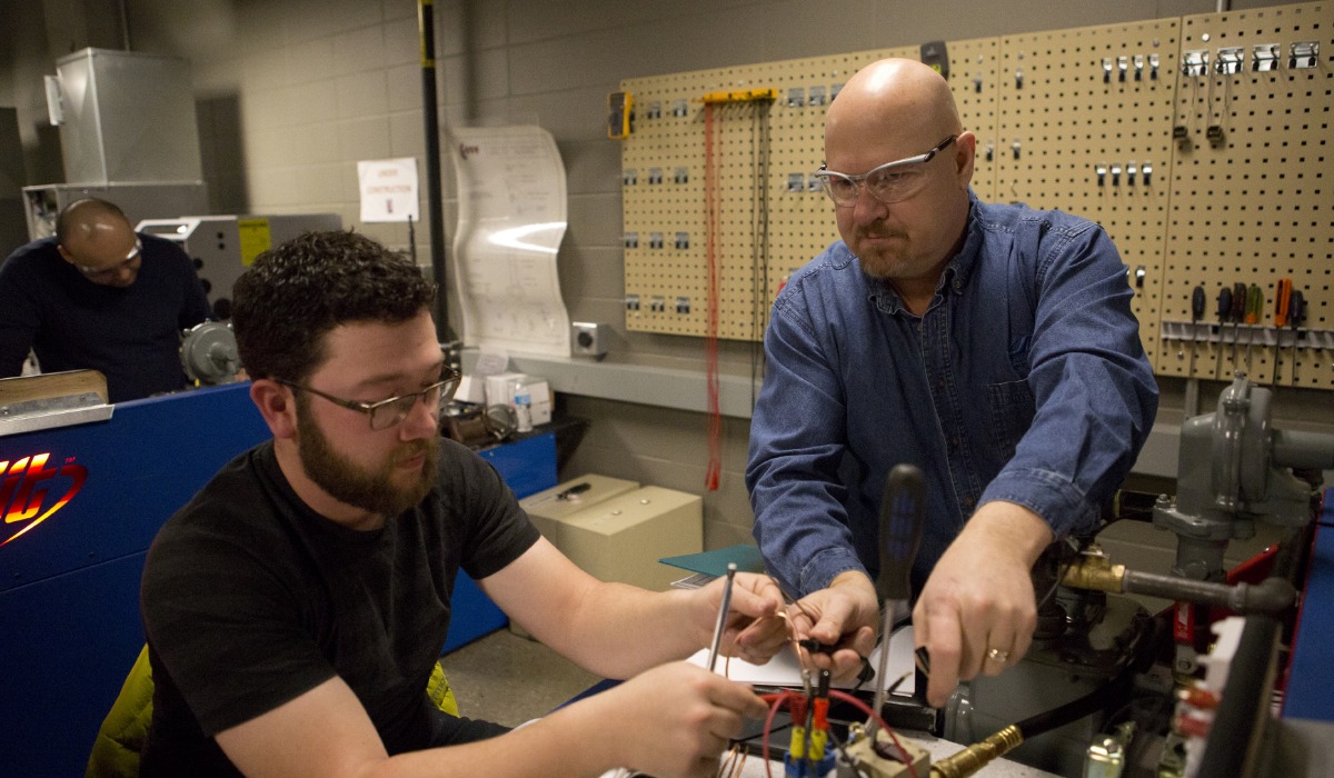 An instructor and a student wearing safety glasses work together on a gasfitting system in a workshop, connecting wires and components while tools hang on a pegboard in the background.