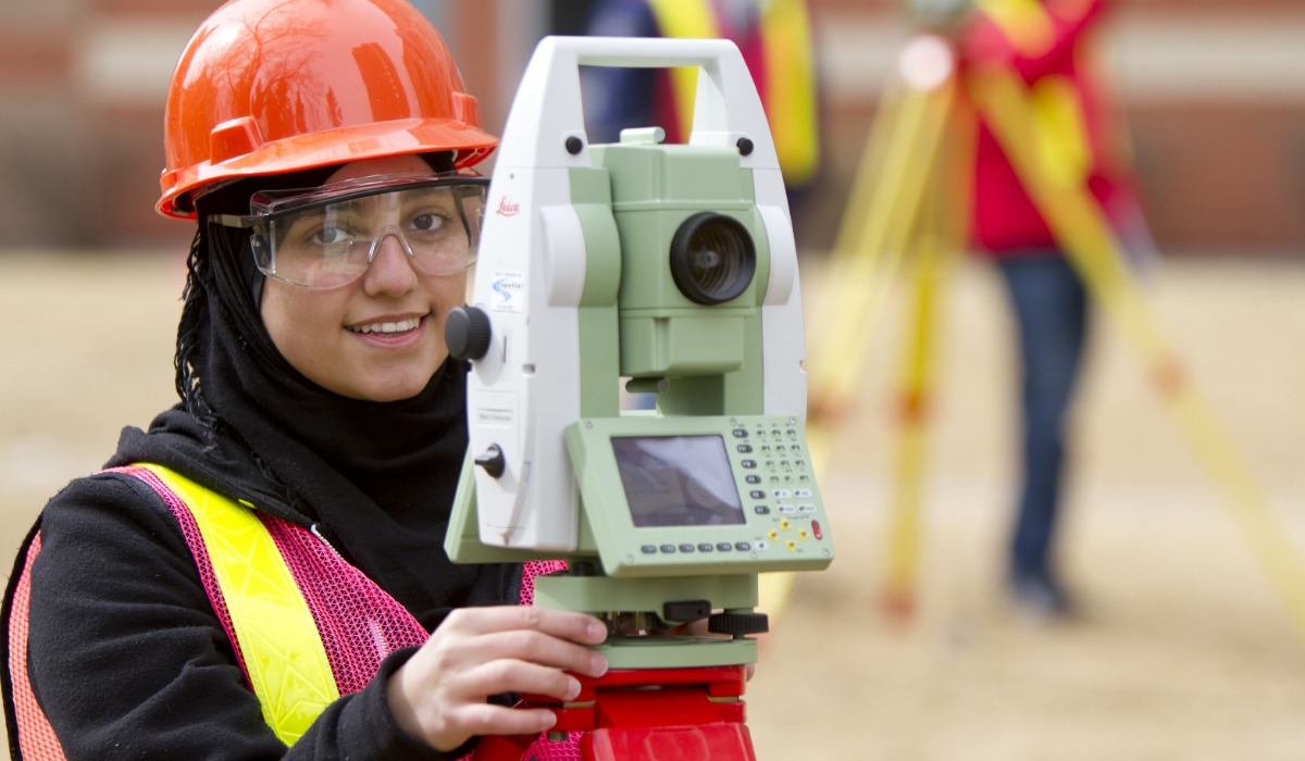 A surveying professional using a total station on a construction site, wearing safety gear and an orange hard hat.