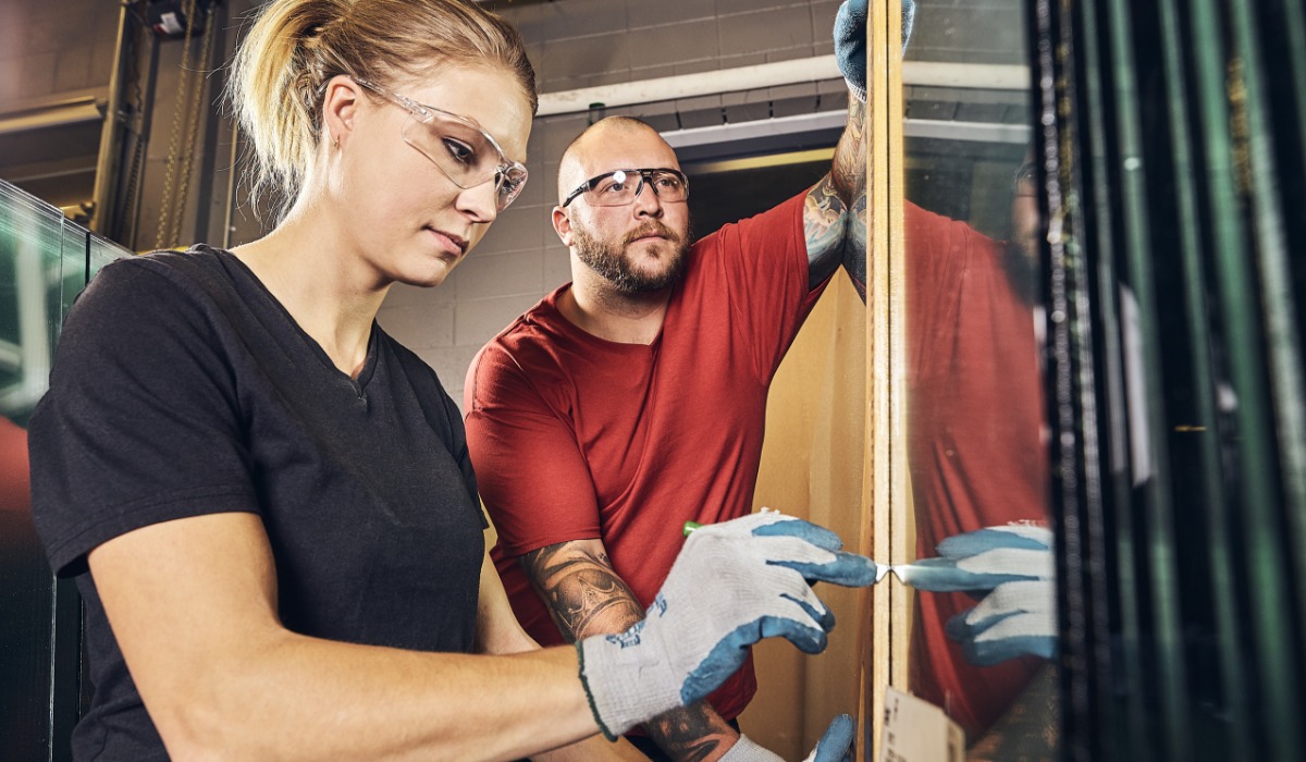 Two apprentices wearing safety glasses and gloves work carefully with a large sheet of glass in a workshop, aligning and securing it into place.