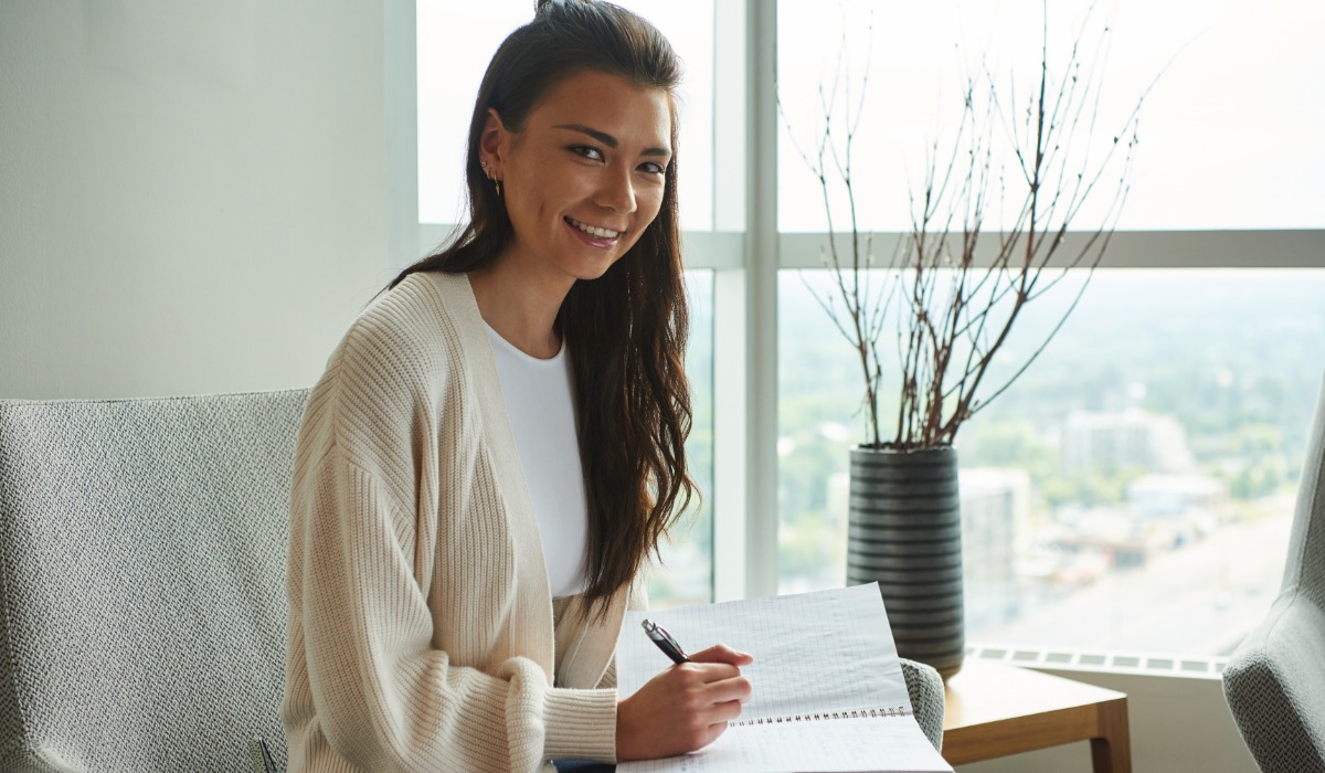 A young woman smiling while writing in a notebook, seated by a large window with a city view.