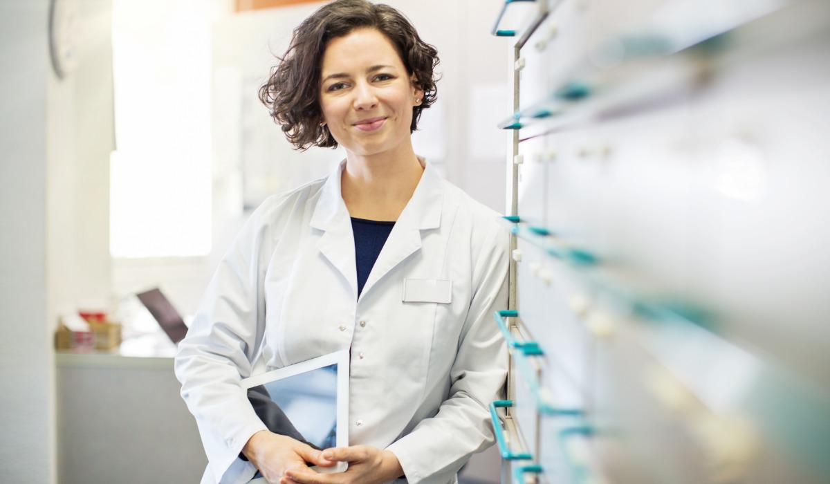 A smiling healthcare professional wearing a lab coat, holding a tablet, and standing in a medical office.