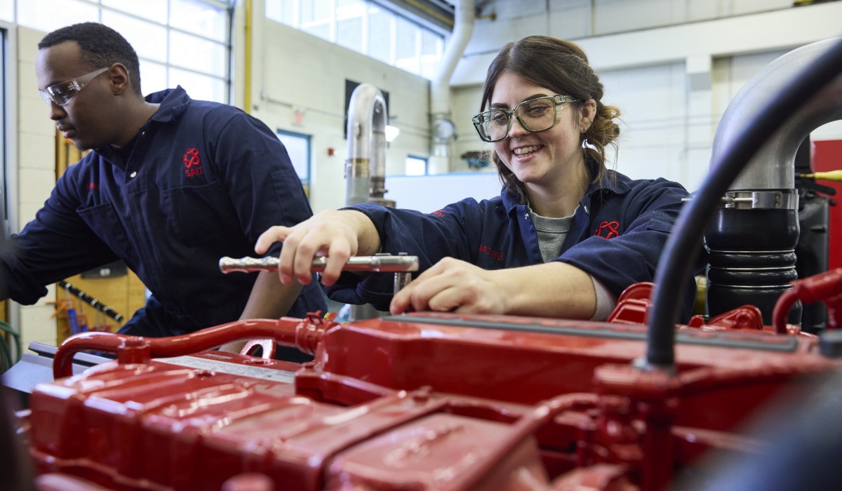 Two apprentices wearing safety glasses and navy coveralls work together on a large red engine in a mechanical workshop, using tools to adjust its components.