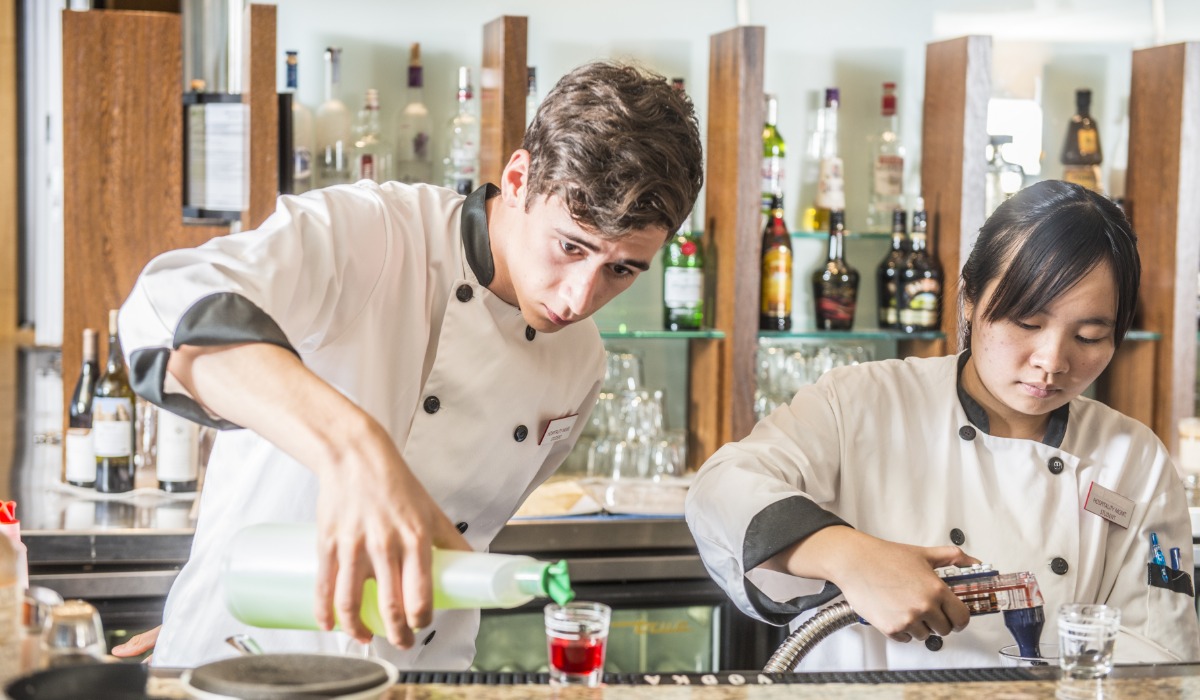 Two bartenders in chef coats are preparing drinks at a bar, with bottles of liquor displayed in the background.