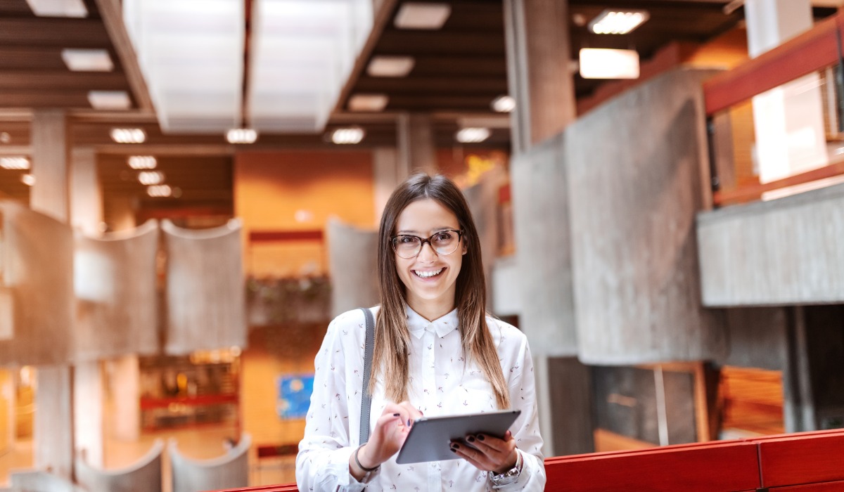 A smiling woman in glasses holding a tablet, set in a modern architectural environment.