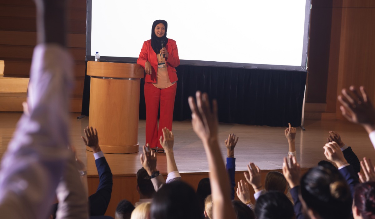 A speaker in a red suit addresses an audience, with numerous hands raised in response.