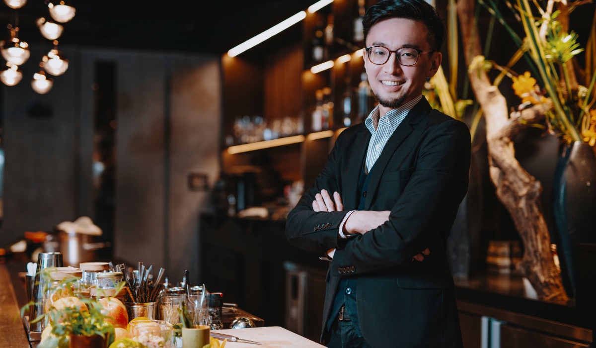 A confident bartender stands with crossed arms in a stylish bar, showcasing a variety of fresh ingredients and cocktails in the background.