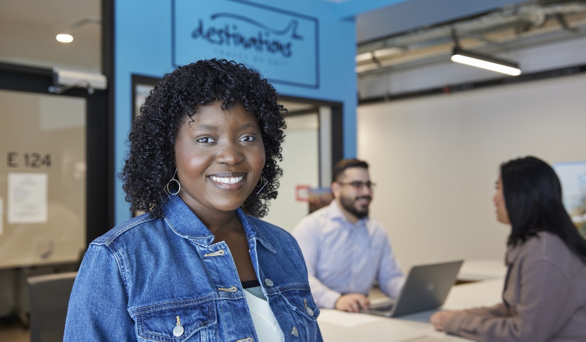 A smiling woman with curly hair stands in an office environment, featuring a desk with two people engaged in discussion. The backdrop includes a sign reading Destinations.