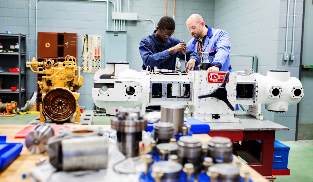 Two apprentices wearing safety glasses work together to assemble or repair a large industrial machine in a workshop, with tools and mechanical parts spread across nearby tables.