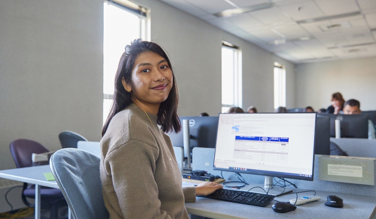 A young woman smiles at the camera while working on a computer in a classroom setting.