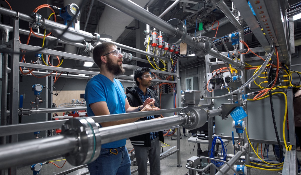 Two apprentices wearing safety glasses examine and discuss a complex network of pipes, sensors, and wiring in an industrial instrumentation and control lab.