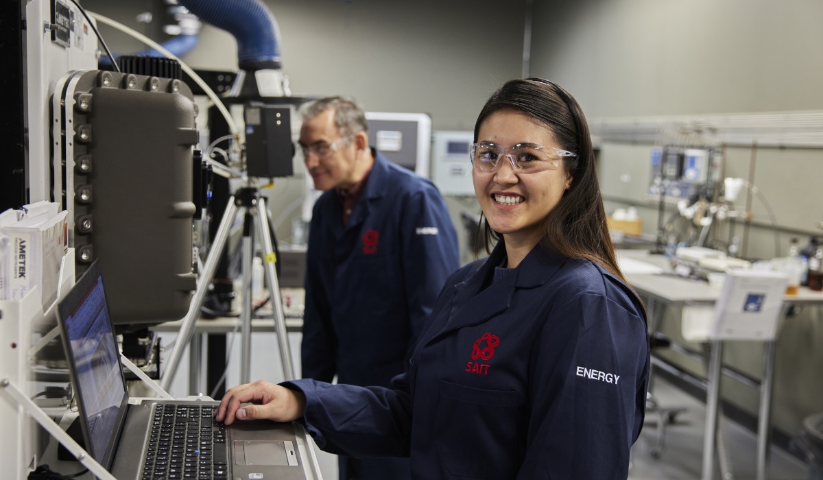 A scientist in a lab coat and safety glasses smiles while using a laptop, with another researcher in the background, showcasing a high-tech research environment.