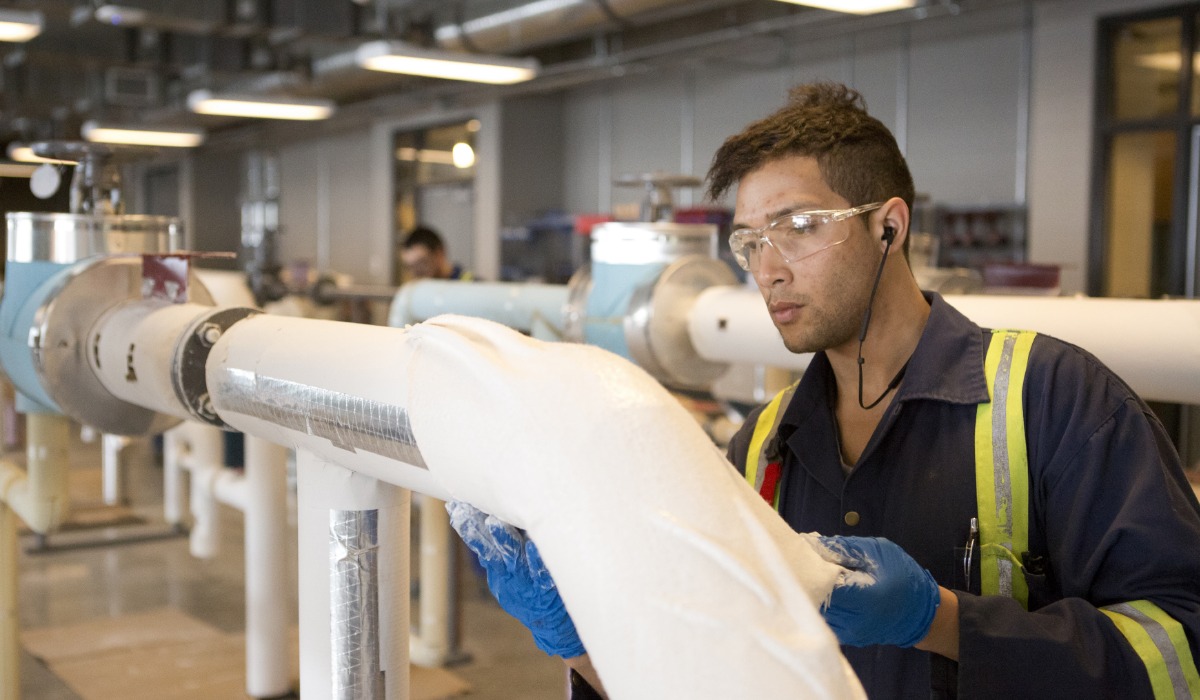 An apprentice wearing safety glasses, gloves, and a reflective uniform applies insulation material to large industrial pipes in a workshop setting.