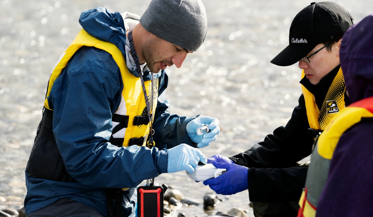 Two researchers, wearing yellow life jackets and gloves, are conducting a field experiment by collecting and analyzing samples at a riverbank.