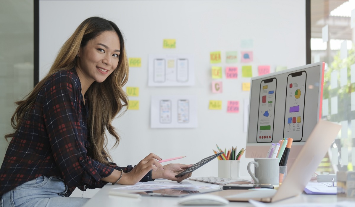 A woman smiling while working on a tablet at a desk filled with design drafts and colorful sticky notes in a modern office environment.