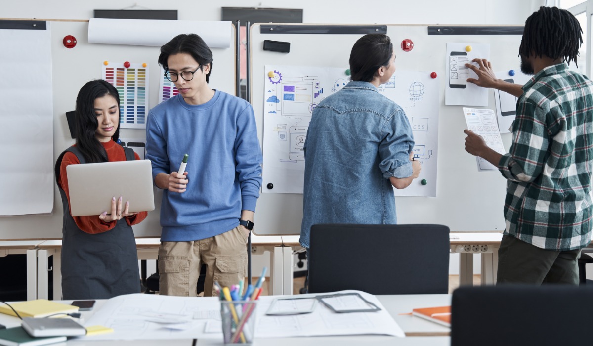 A group of four diverse individuals collaborating in a workspace, actively discussing ideas and working on a project presentation displayed on boards.