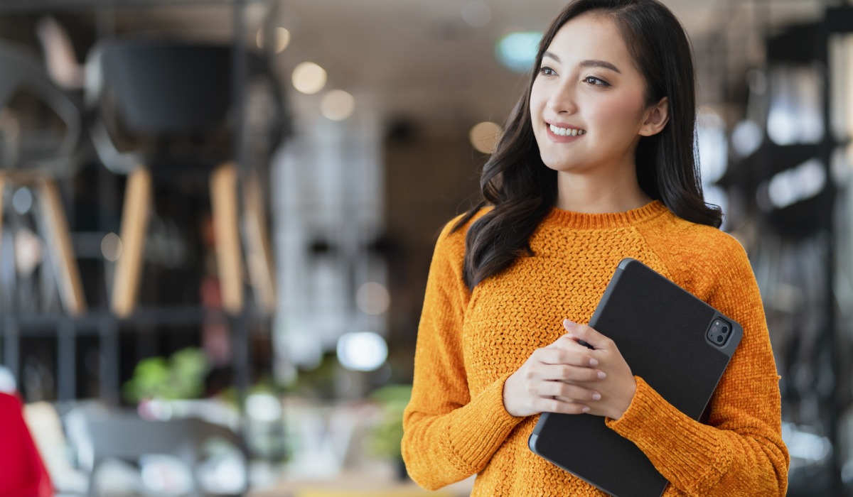 A young woman in a cozy orange sweater holds a tablet while smiling, set against a modern café backdrop.
