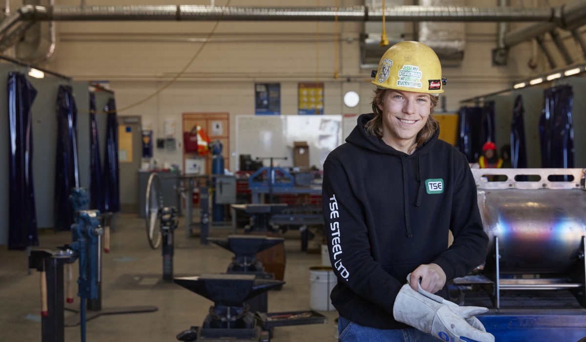 A smiling ironworker wearing a yellow hard hat, gloves, and a black hoodie stands in a workshop with metalworking tools and equipment in the background.
