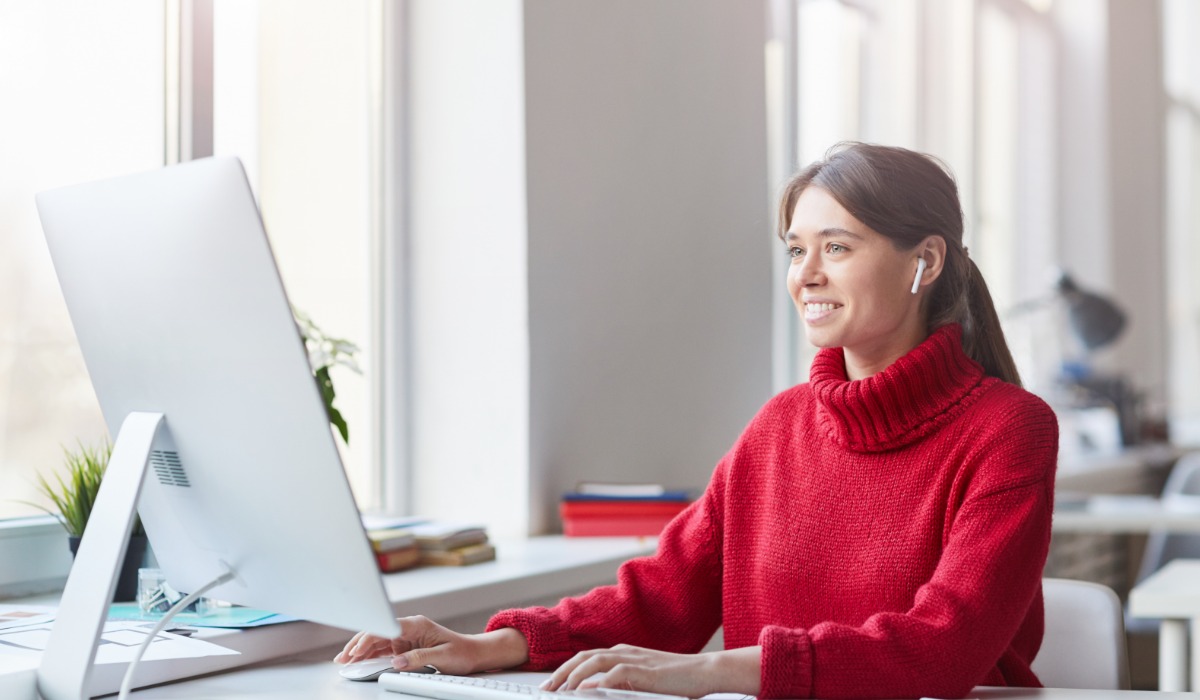 A woman wearing a red sweater is smiling while working on a computer in a well-lit office.