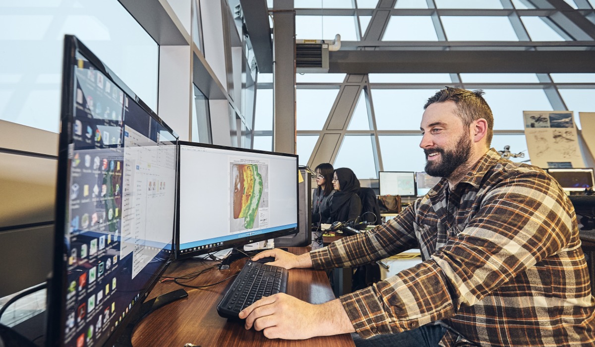 A man working at a desk with dual monitors, focused on a digital project, in a modern office environment.