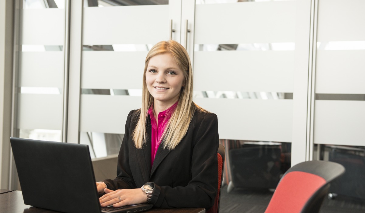 A professional woman seated at a table with a laptop, wearing business attire, and smiling in a modern office environment.
