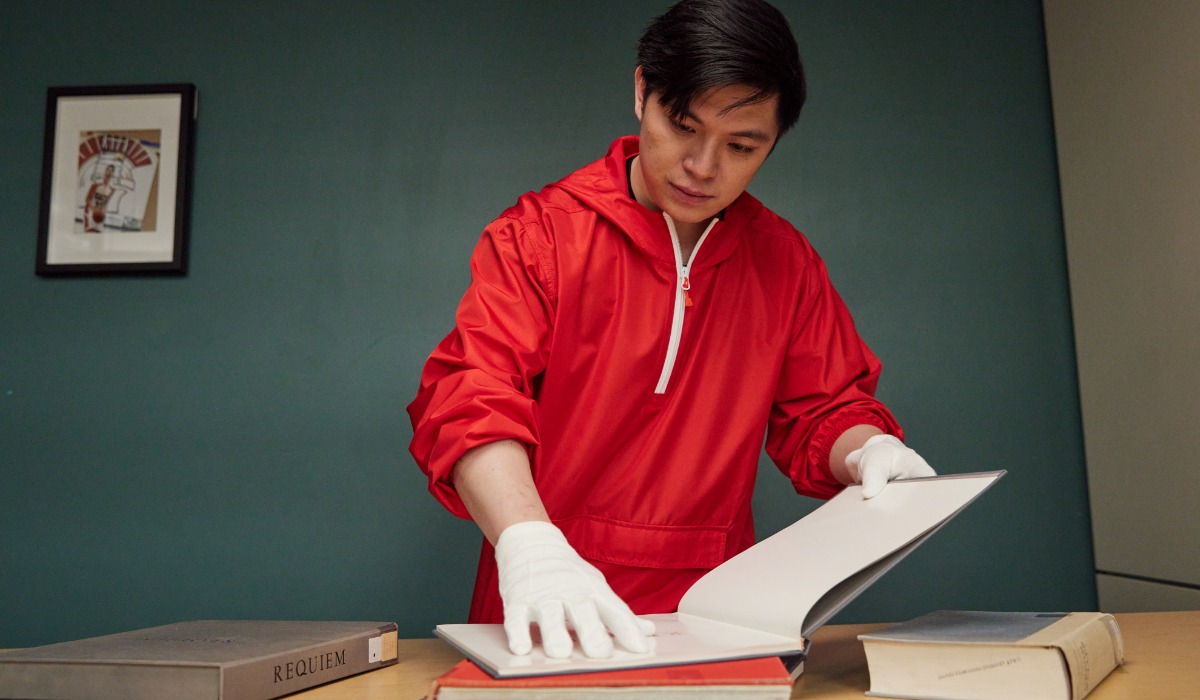 A man in a red jacket examines a book while wearing white gloves, suggesting care in handling valuable items.