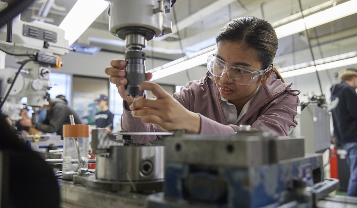 An apprentice wearing safety glasses operates a milling machine in a workshop, adjusting metal components with precision while other students work in the background.