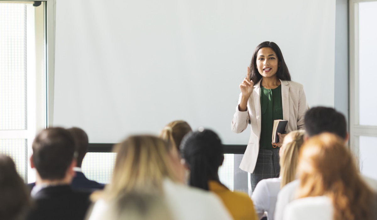 A professional woman stands in front of an audience while presenting, holding a book and gesturing with her hand.