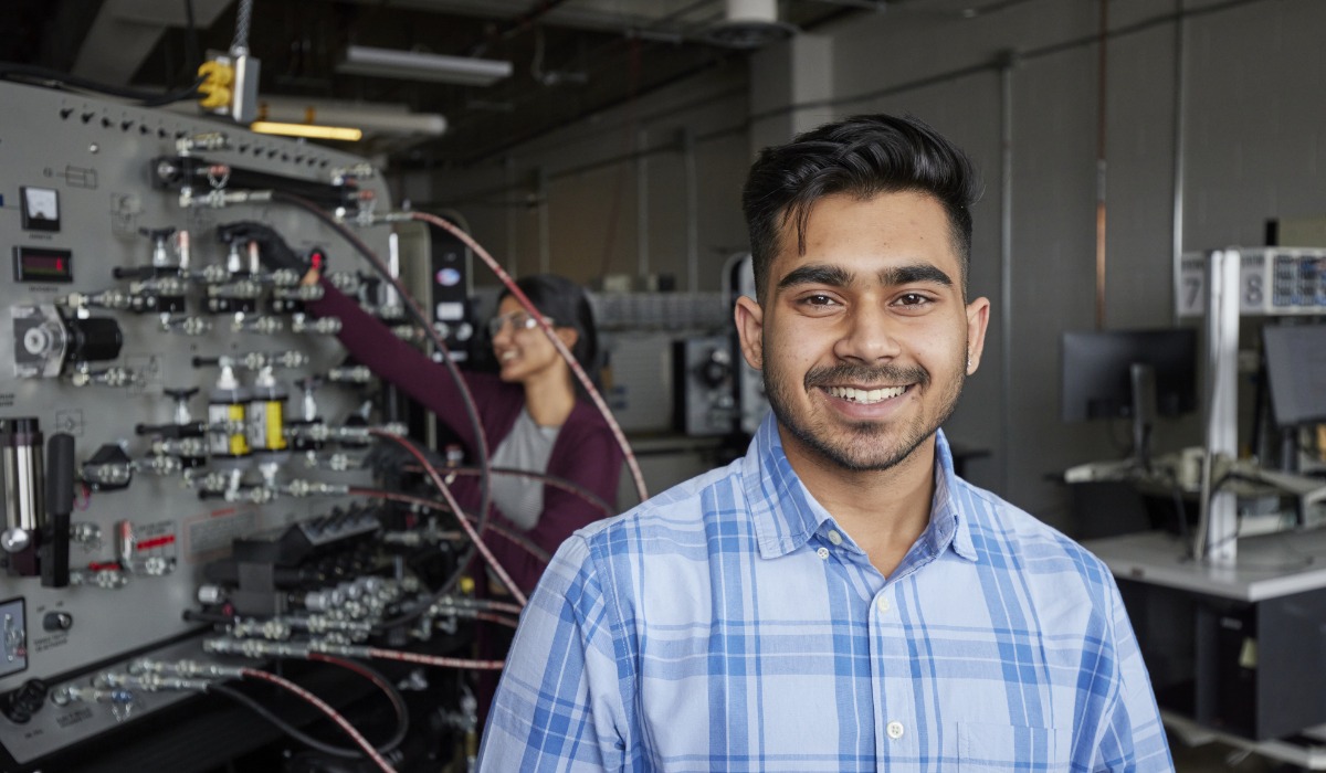 A smiling student stands in a lab with scientific equipment in the background, while a fellow student adjusts the apparatus.