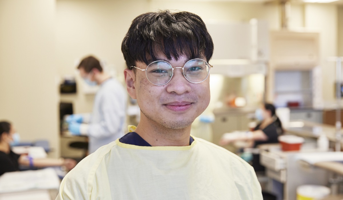 A young male healthcare professional wearing glasses and a yellow medical gown, smiling in a clinical setting.