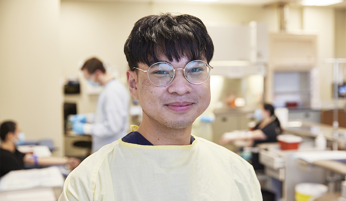 A young male healthcare professional wearing glasses and a yellow medical gown, smiling in a clinical setting.