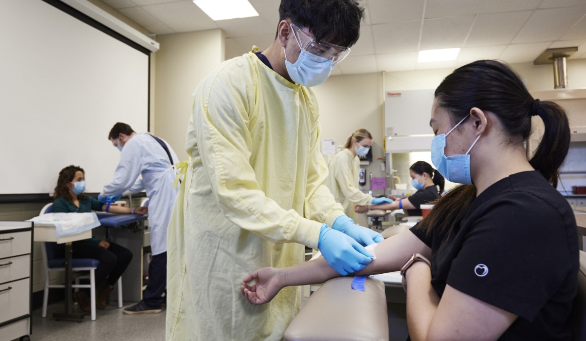 Healthcare professionals in personal protective equipment are conducting a blood draw on a patient in a clinical training environment.