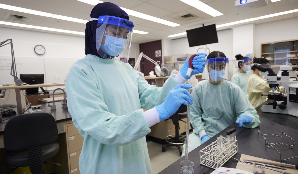 Students in a science laboratory wearing protective gowns, gloves, face shields, and masks. One student in the foreground is using a pipette to transfer liquid into a test tube, while others observe and work with microscopes in the background.
