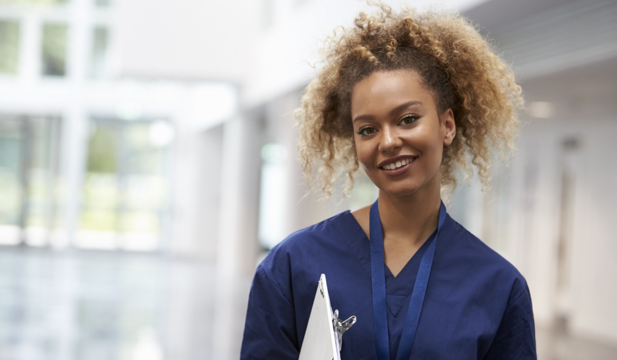 Smiling healthcare professional in blue scrubs holding a clipboard in a bright, modern medical facility.