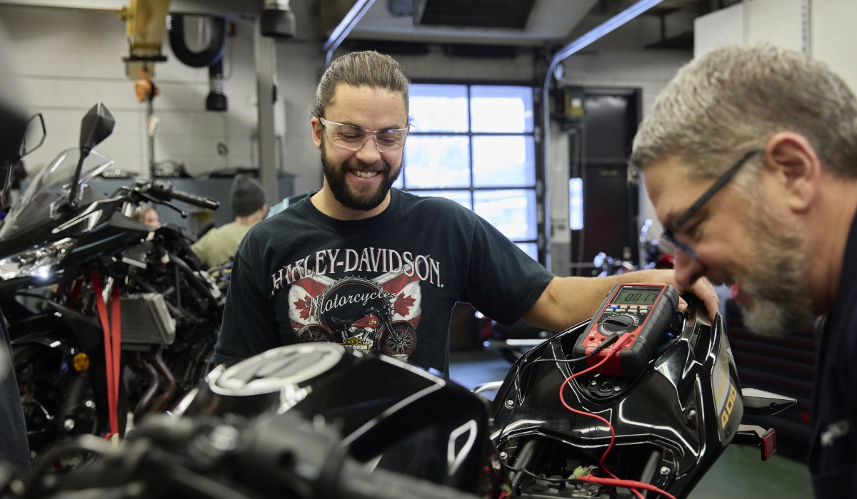Two apprentices in a motorcycle workshop smile while using diagnostic equipment to test a motorcycle’s electrical system, surrounded by bikes and tools.